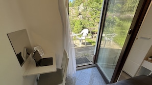 Una habitació amb llit, escriptori amb portàtil, cadira i una porta de vidre corredissa que dona a una zona exterior amb cadires i vegetació.