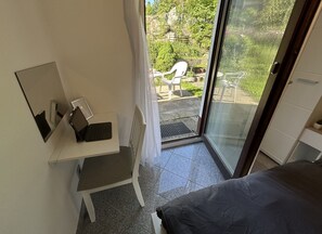 Een kamer met een bed, een bureau met een laptop, een stoel en een glazen schuifdeur die leidt naar een buitenruimte met stoelen en groen.
