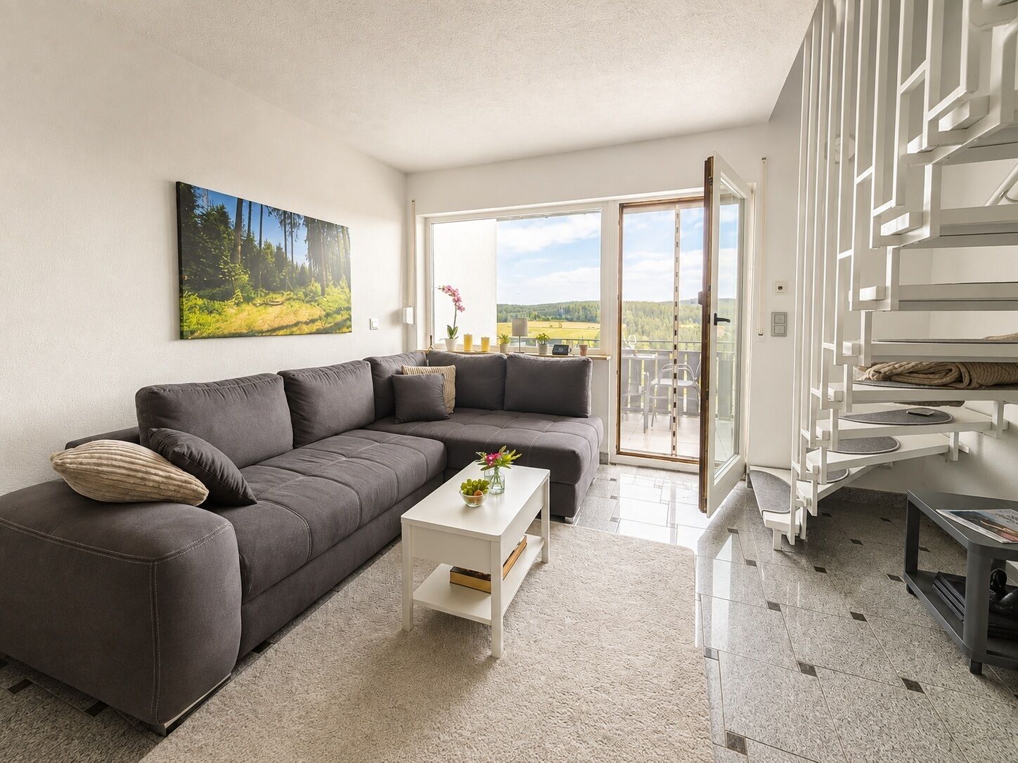 Een moderne woonkamer met een grijze bank, een witte salontafel en een balkon met uitzicht op het platteland.