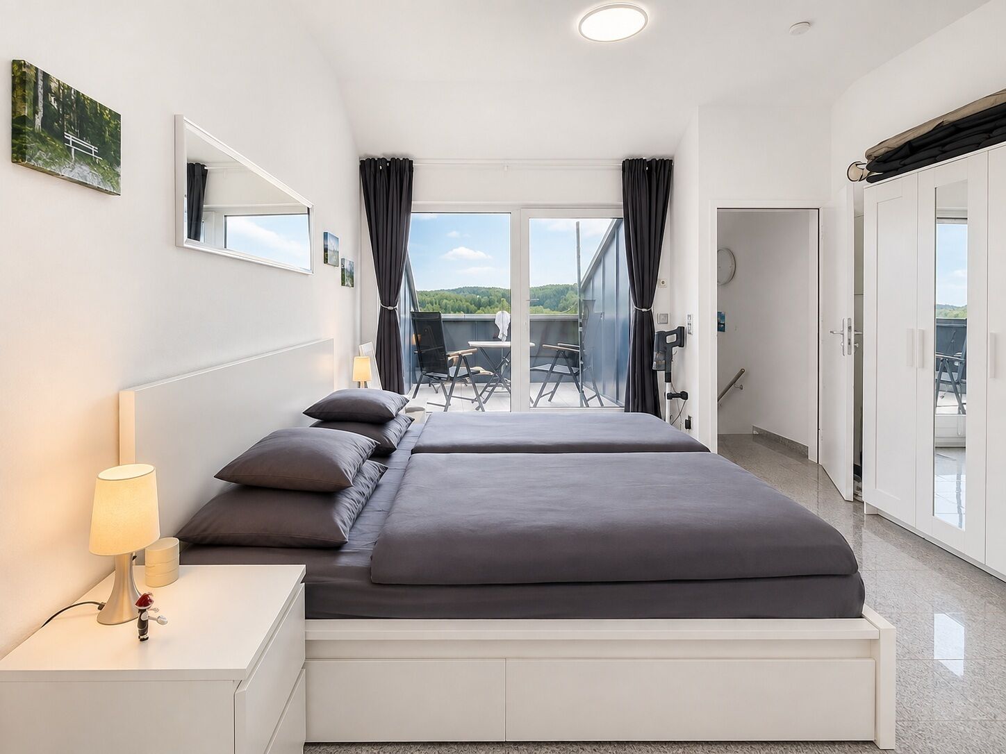 A modern hotel room with a large bed, a bedside table with a lamp, a view of the outdoors, and a balcony with chairs.