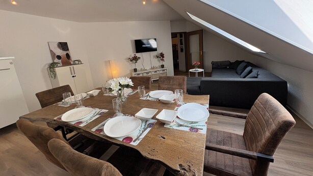 Un comedor con una mesa dispuesta para cuatro personas, un sofá y un televisor montado en la pared.