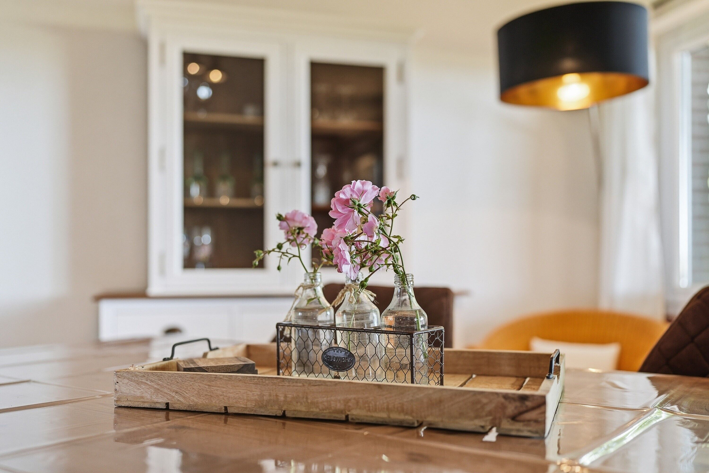 En bakke med glasflasker fyldt med lyserøde blomster, en træbakke og et metalstativ med et skilt.