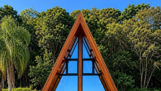 Una estructura de fusta triangular amb façana de vidre, envoltada de vegetació abundant.