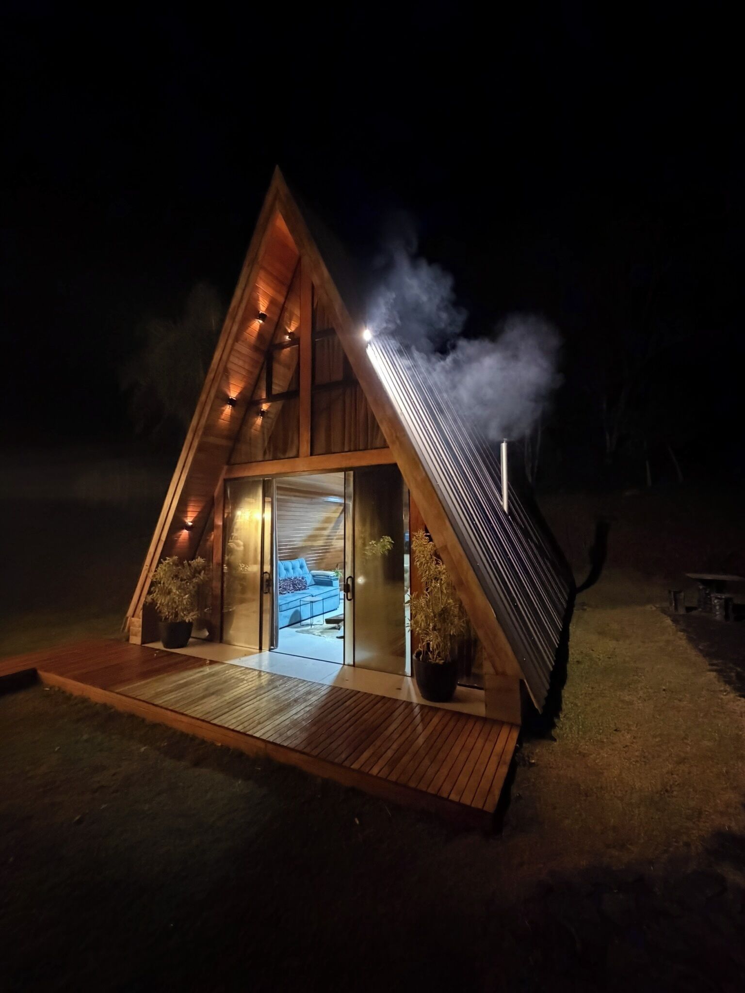 A triangular wooden structure with a glass entrance, a blue sofa inside, and a smoke stack emitting smoke.