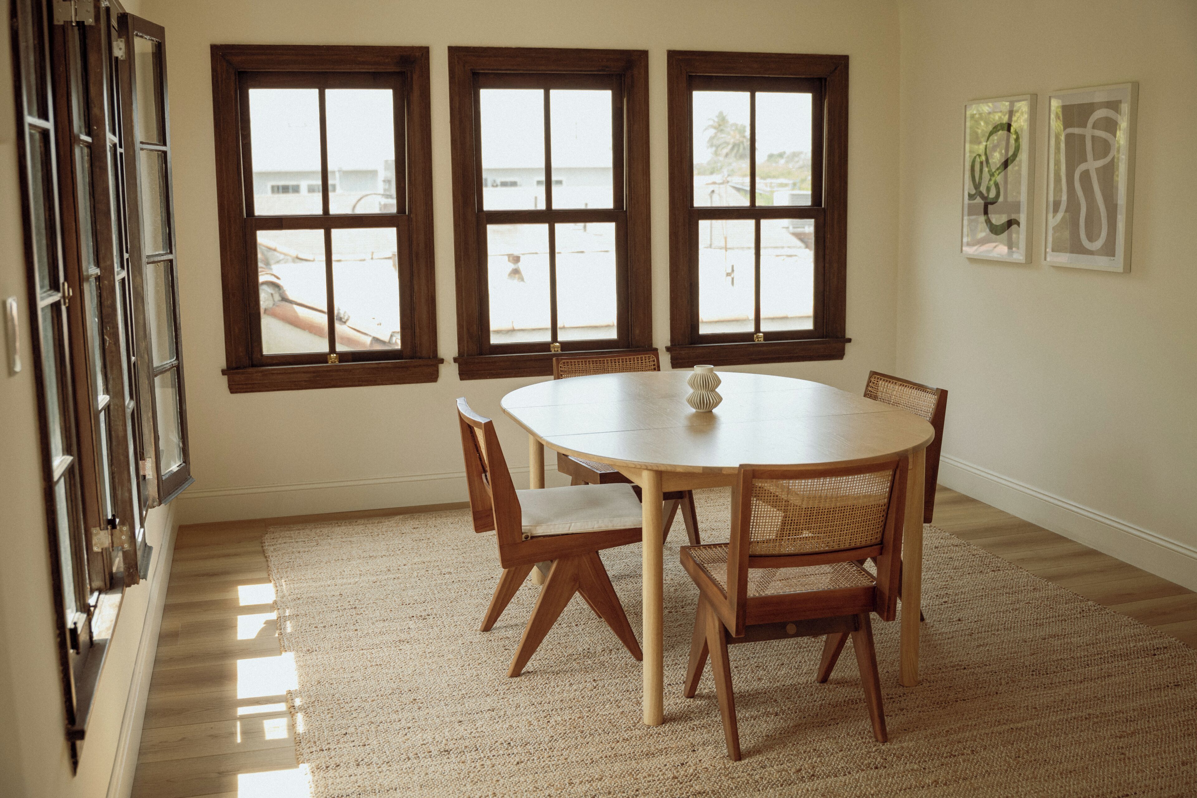 A dining area with a round table and four chairs, three windows, and a rug on the floor.