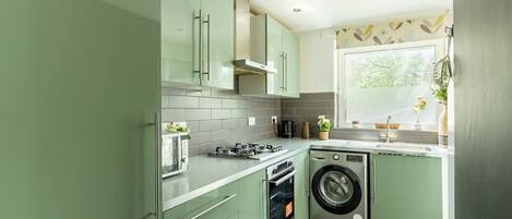 Cucina con mobili verdi, piano cottura a gas, lavatrice e finestra vista su spazio verde.