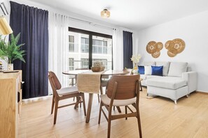 A modern living room with a dining area, a white sofa, and a large window with curtains. - Modern 1BR w/2 Pools + Balcony Near Beach & Dining (Punta Cana)
