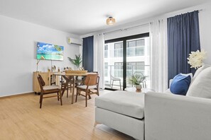 A modern living room with a sofa, dining table, chairs, and a large window with curtains. - Modern 1BR w/2 Pools + Balcony Near Beach & Dining (Punta Cana)