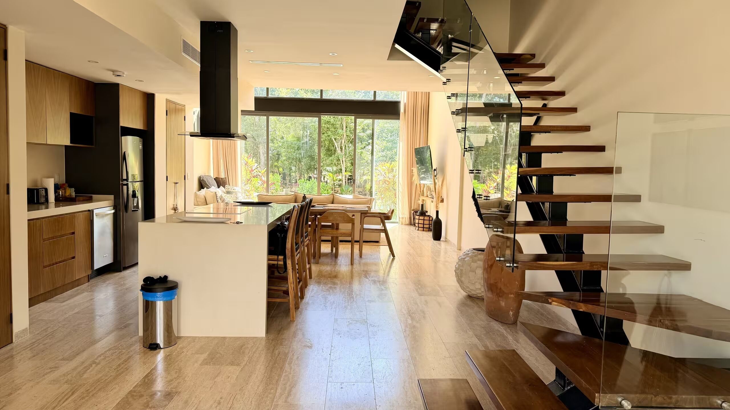 Una cocina moderna con una isla central, un área de comedor con una mesa de vidrio y una escalera con barandales de vidrio.