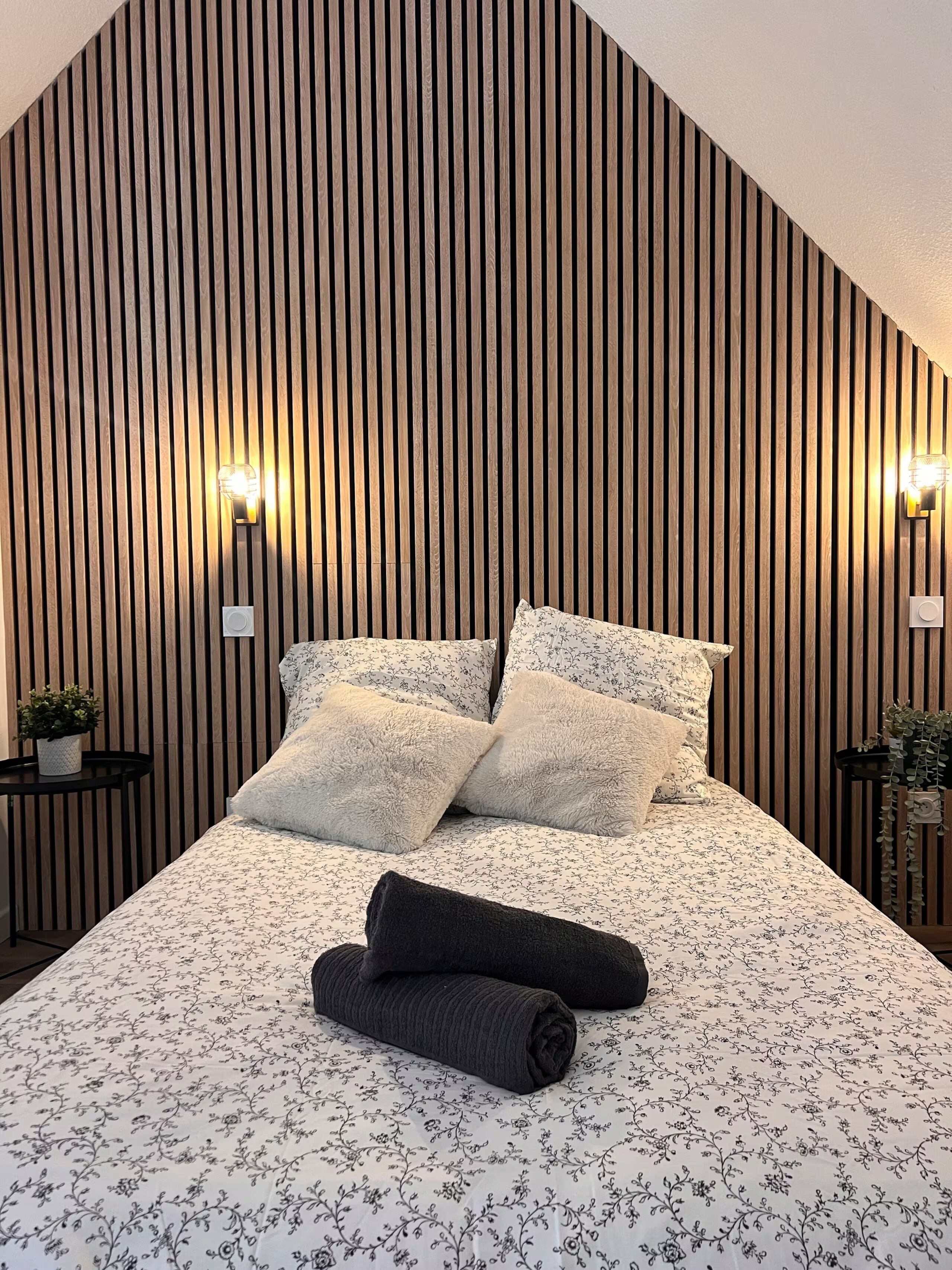 Een slaapkamer met een wand van houten latten, twee nachtkastjes met lampen, een bed met bloemenbedrukking en een opgerolde handdoek op het bed.