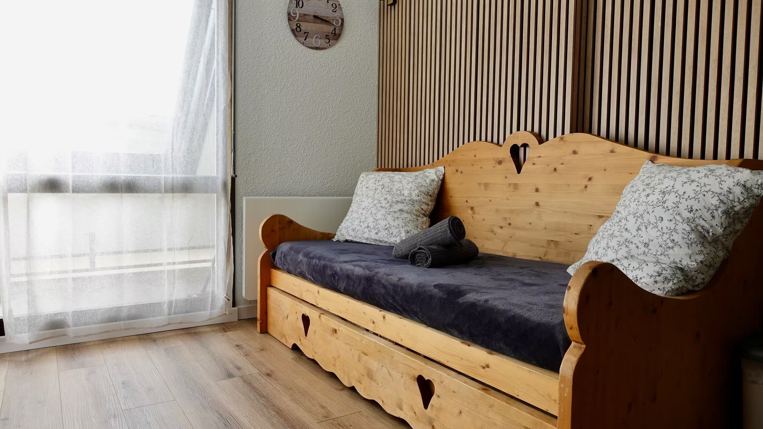 Un canapé en bois avec un coussin foncé, deux coussins à motifs et une horloge murale.
