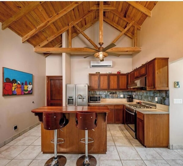 A kitchen with wooden cabinets, a central island with stools, a ceiling fan, and a painting on the wall. - Come hang out for a while! (Sandy Bay)