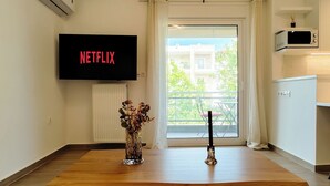 A modern living room with a wooden table, a vase with flowers, a flat-screen TV displaying 'NETFLIX,' and a view of the outside balcony. - Modern Coastal Escape in Paleo Faliro (Paleo Faliro)