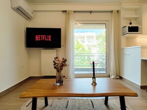 A modern living room with a wooden table, a vase with flowers, a flat-screen TV displaying 'NETFLIX,' and a view of the outside balcony. - Modern Coastal Escape in Paleo Faliro (Paleo Faliro)