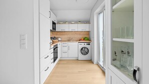 Une cuisine moderne avec des meubles blancs, un plan de travail en bois, un lave-linge et un meuble vitré contenant des bouteilles.