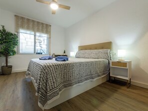 Ein Schlafzimmer mit Bett, Deckenventilator, Fenster mit Jalousien, Nachttisch mit Lampe und einer Zimmerpflanze.