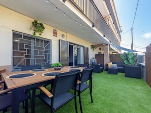 Um pátio externo com mesa e cadeiras de madeira, uma pequena área de jardim com plantas e um edifício com varanda.