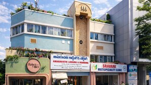 Rakennuksessa on kyltti, jossa lukee »Amaravathi Lodging & Boarding» ja »Sravani Multispecialty Clinic». Sen julkisivu on sinivalkoinen, ja alakerrassa on pieni myymälä nimeltä »Baker's Mill». Lisäksi rakennuksen edessä on muutamia parkkeerattuja moottoriskoottereita.