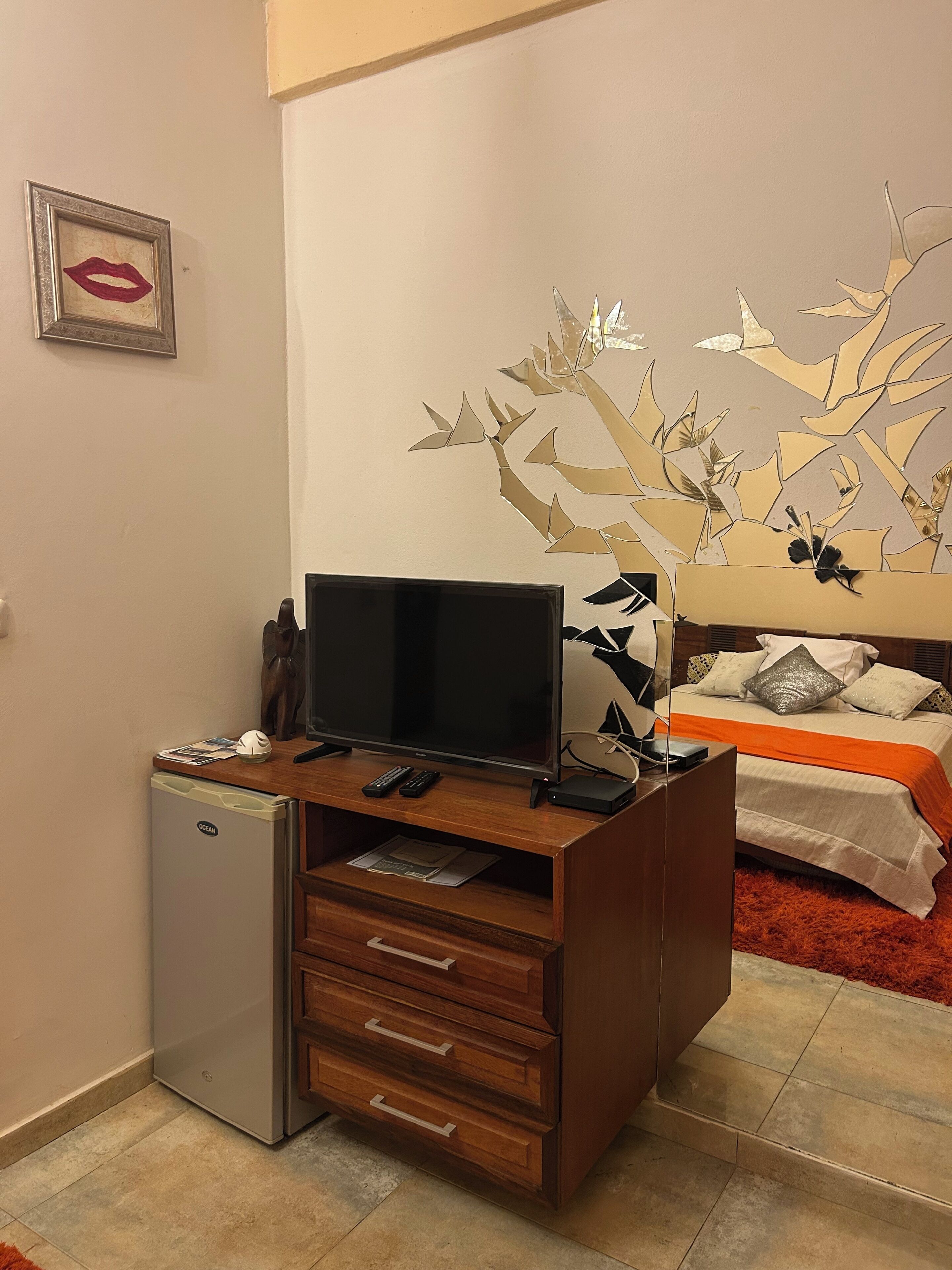Een knus ingerichte kamer met een kleine koelkast, een houten bureau met een flatscreen televisie, een bed met een rode vloerkleed en een decoratief kunstwerk aan de muur.