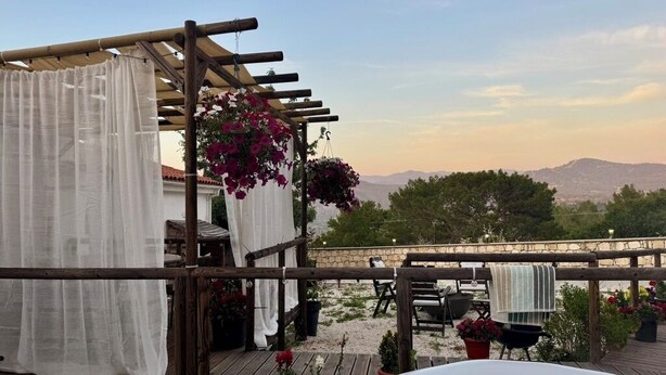 Ein Whirlpool, eine Holzterrasse, eine Pergola mit hängenden Blumen und Blick auf ferne Berge.