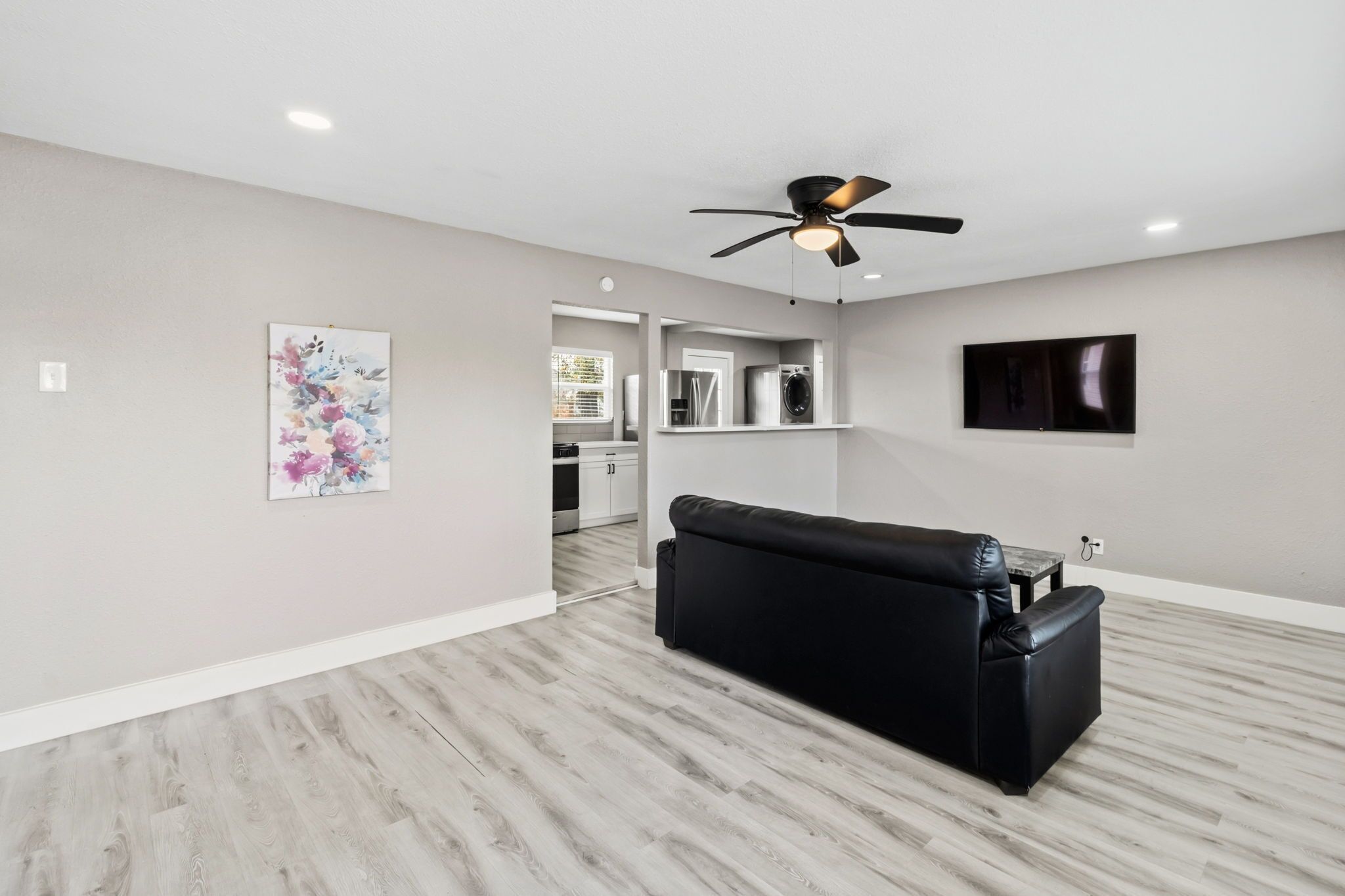 A modern living room with a black leather sofa, a ceiling fan, a flat-screen TV, and a kitchen area in the background.