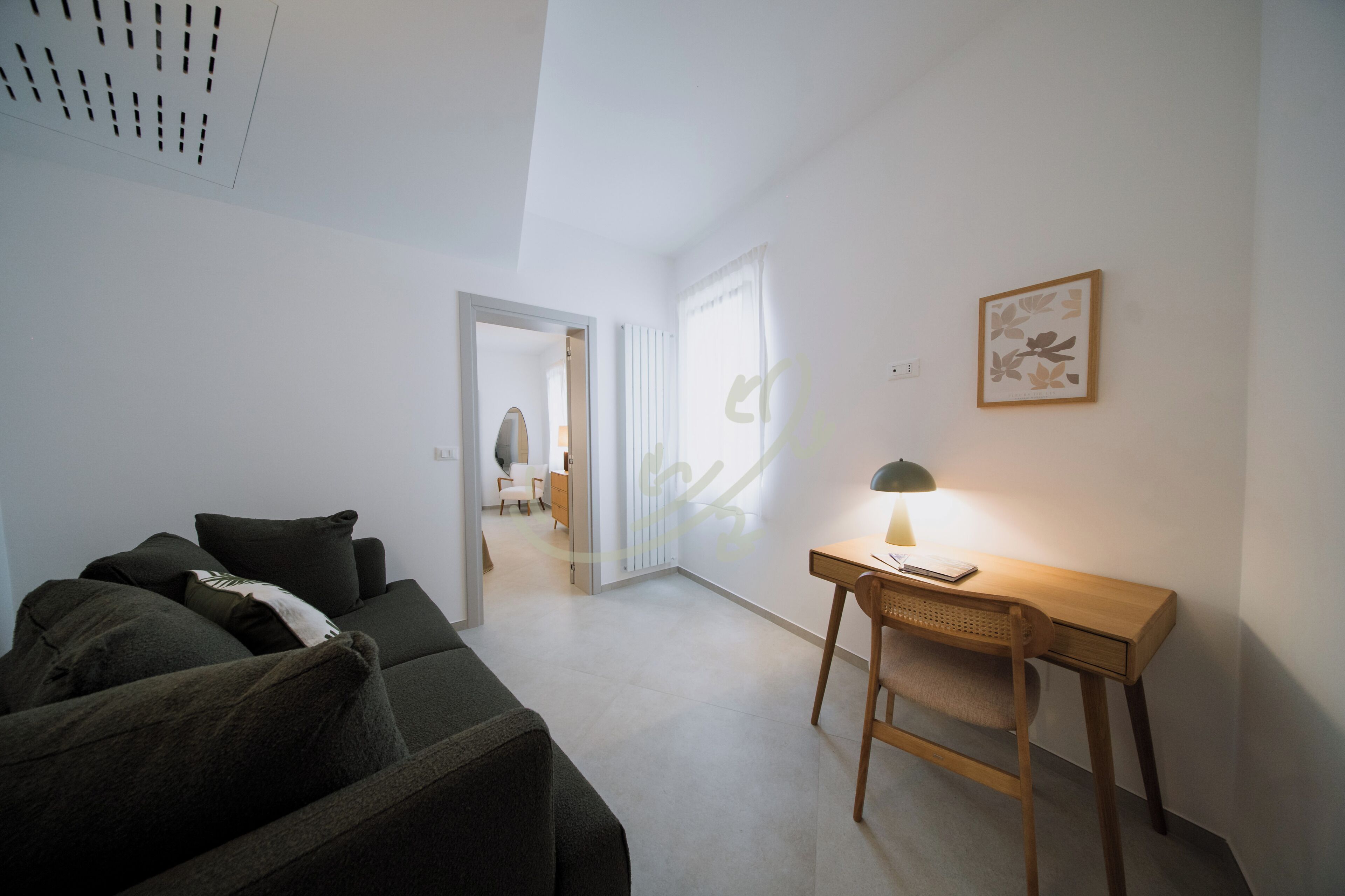 Et værelse med en sofa, et skrivebord med en lampe, et billede i ramme på væggen og en dør til et andet rum.
