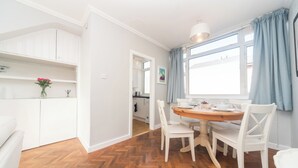A dining area with a wooden table and white chairs, a kitchen in the background, and a window with blue curtains. - Fulham 2BR Flat | Clean, Quiet & Spacious - Pass the Keys (London)