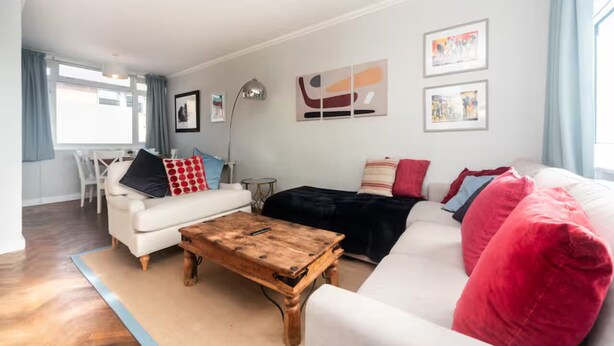 A living room with a white sofa, a wooden coffee table, and a black sofa with red cushions. There are framed pictures on the wall and a lamp on a side table. - Fulham 2BR Flat | Clean, Quiet & Spacious - Pass the Keys (London)