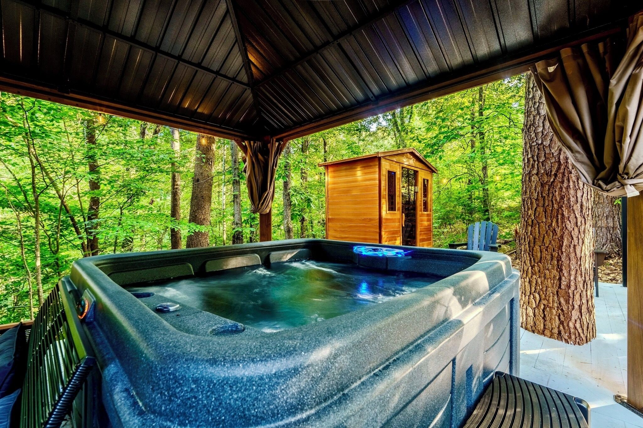 A hot tub with a view of a forest, a wooden cabin, and a tree trunk.