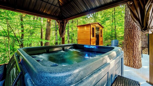 A hot tub with a view of a forest, a wooden cabin, and a tree trunk. - Luxury Couples Cabin | Hot Tub + Sauna + Theater (Sevierville)