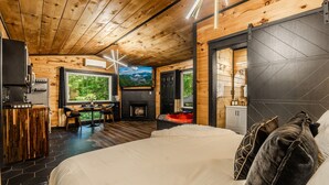 A wooden cabin interior with a bed, a dining area, a fireplace, and a large window. - Luxury Couples Cabin | Hot Tub + Sauna + Theater (Sevierville)