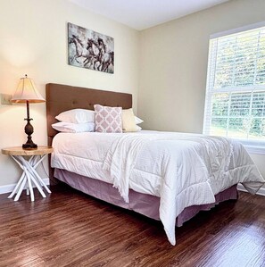 Quarto com cama, mesa de cabeceira, luminária, quadro e janela com persianas.