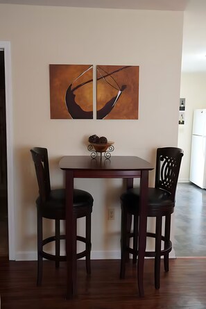 Un comedor con una mesa y dos sillas, una obra de arte en la pared y un refrigerador al fondo.