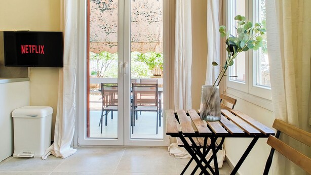 A room with a sliding glass door leading to an outdoor area, a table with benches, a trash bin, and a TV mounted on the wall displaying 'NETFLIX'. - Hidden Garden Home in Glyfada Center (Glifada)