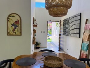Ein Essbereich mit einem Holztisch, schwarzen Stühlen, gewobenen Tischsets, einem Korbkissen und einem Wandbild. Ein Durchgang führt nach draußen.