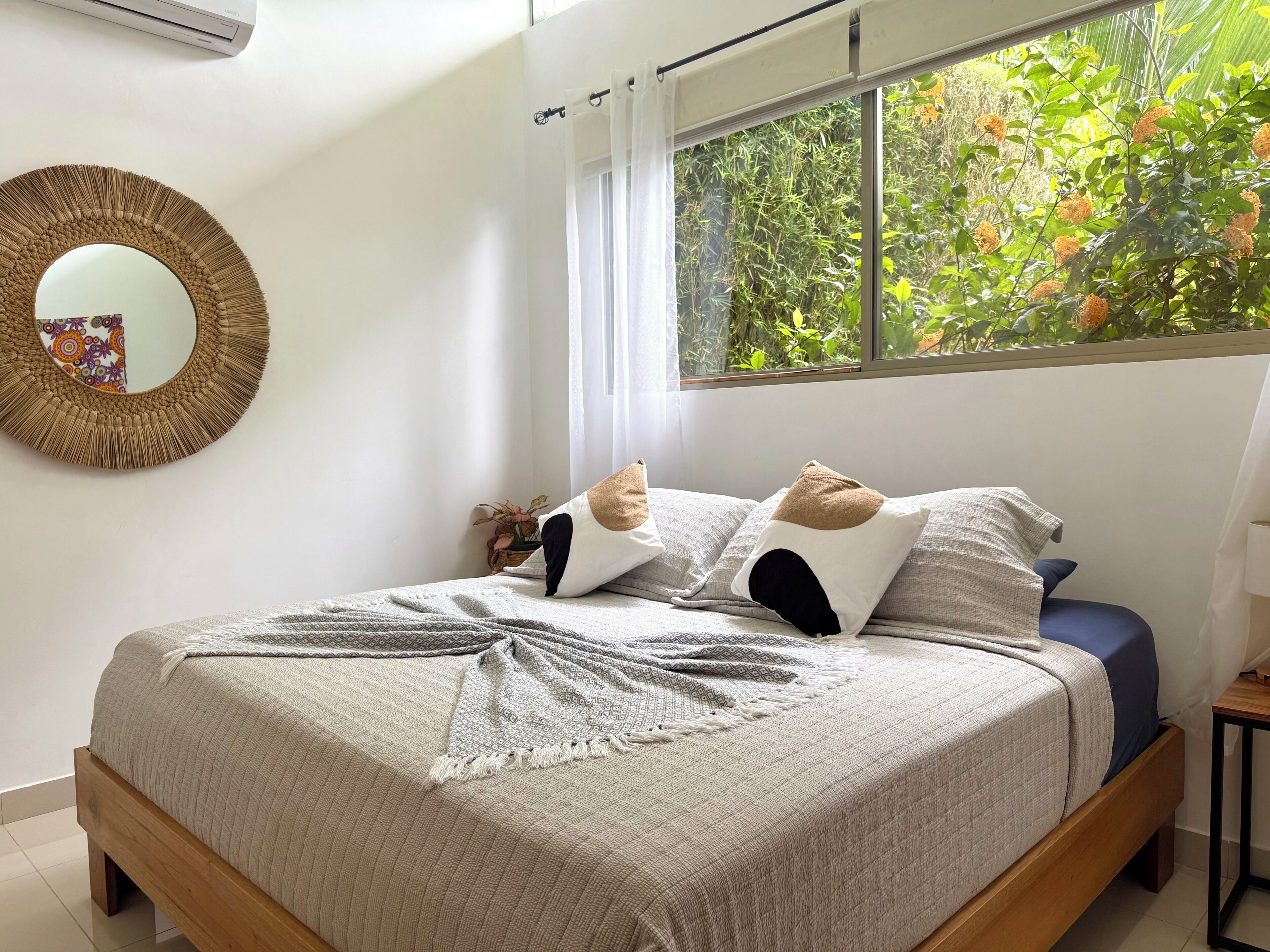 A bedroom with a bed, a mirror, a window with a view of greenery, and a small table with a lamp.