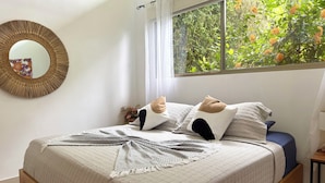 Un dormitorio con cama, espejo, ventana con vista a la vegetación y una mesita de noche con lámpara.