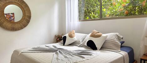 A bedroom with a bed, a mirror, a window with a view of greenery, and a small table with a lamp.