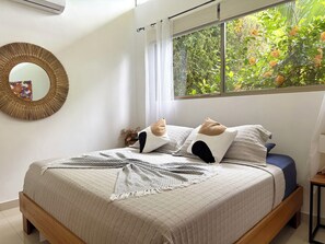 A bedroom with a bed, a mirror, a window with a view of greenery, and a small table with a lamp. - Bali Style • Villa • Near Whales Tail Beach (Uvita)