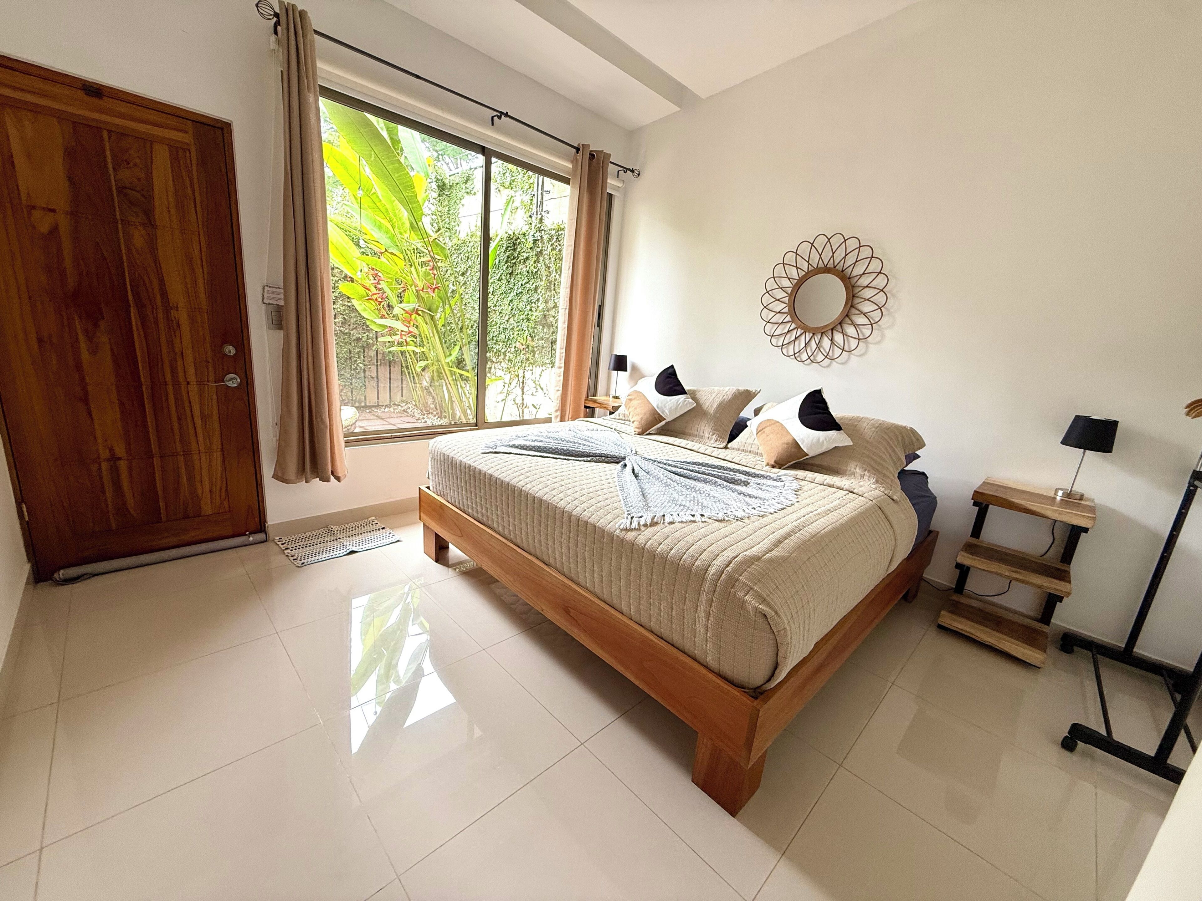 A bedroom with a wooden bed, a bedside table, a mirror, and a view of greenery through the window.