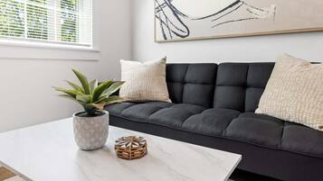 Un salón moderno con un sofá oscuro, una mesa de centro de mármol, una planta en maceta, una cesta tejida y un cuadro abstracto grande en la pared.
