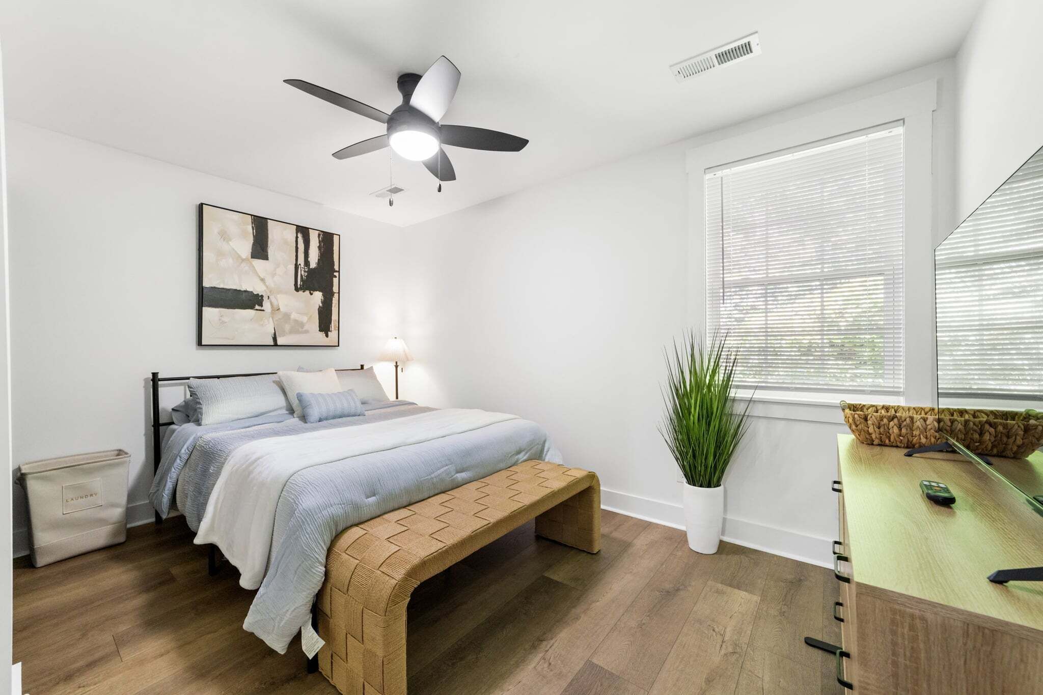 Un dormitorio con cama, ventilador de techo, ventana con persianas, mesita de noche con una planta y un banco.