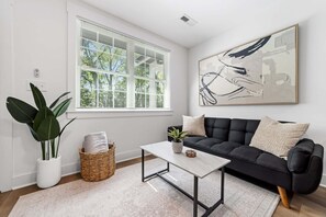 A modern living room with a black sofa, a white coffee table, a large abstract painting, a potted plant, and a window with blinds. - Modern 1BD Condo - Downtown Walkable to F&M Bank Arena (Clarksville)