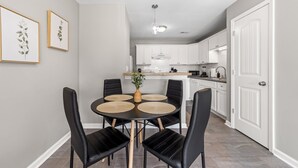 Uma cozinha moderna com armários brancos, uma área de jantar com mesa redonda e cadeiras pretas, e impressões botânicas emolduradas na parede.