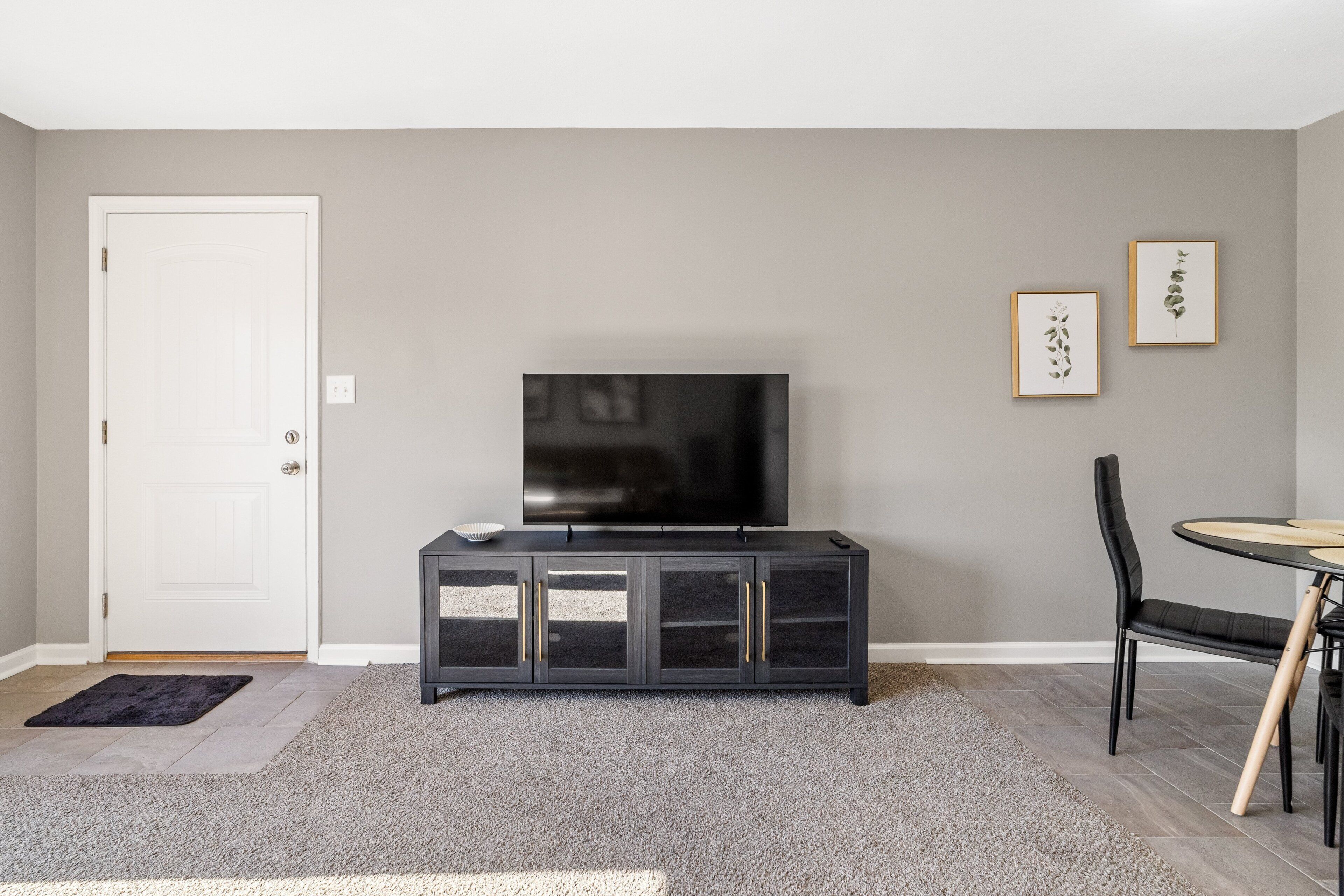 A living room with a black entertainment unit, a flat-screen TV, a dining table with chairs, and two framed botanical prints on the wall.