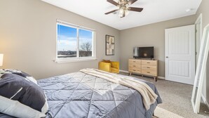 Quarto com cama, ventoinha no teto, cômoda, televisão e janela com vista.