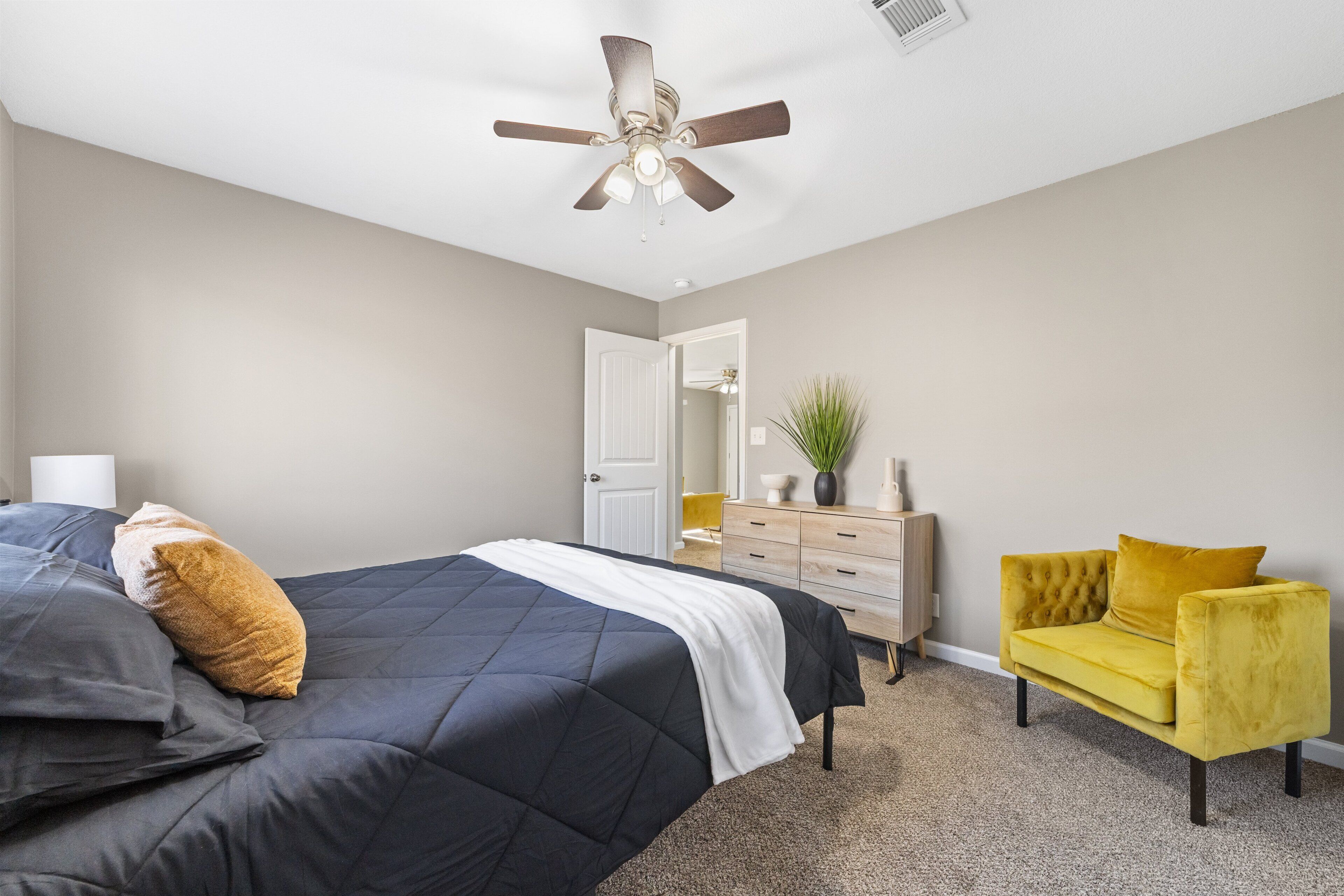 A bedroom with a bed, a yellow armchair, a dresser, a ceiling fan, and a door leading to another room.