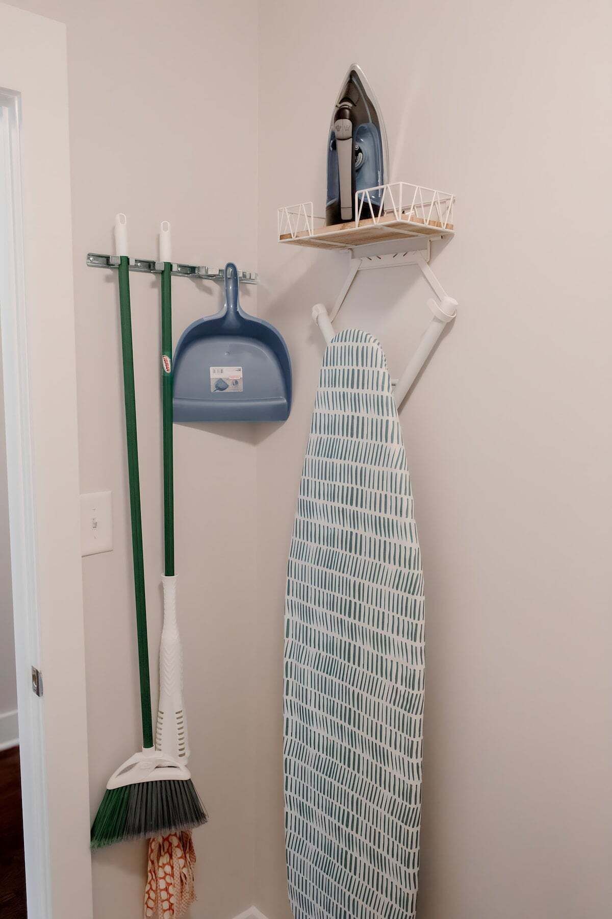 A bathroom with a hanging rack, a blue pan, a green broom, and a patterned ironing board.