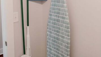 A bathroom with a hanging rack, a blue pan, a green broom, and a patterned ironing board.