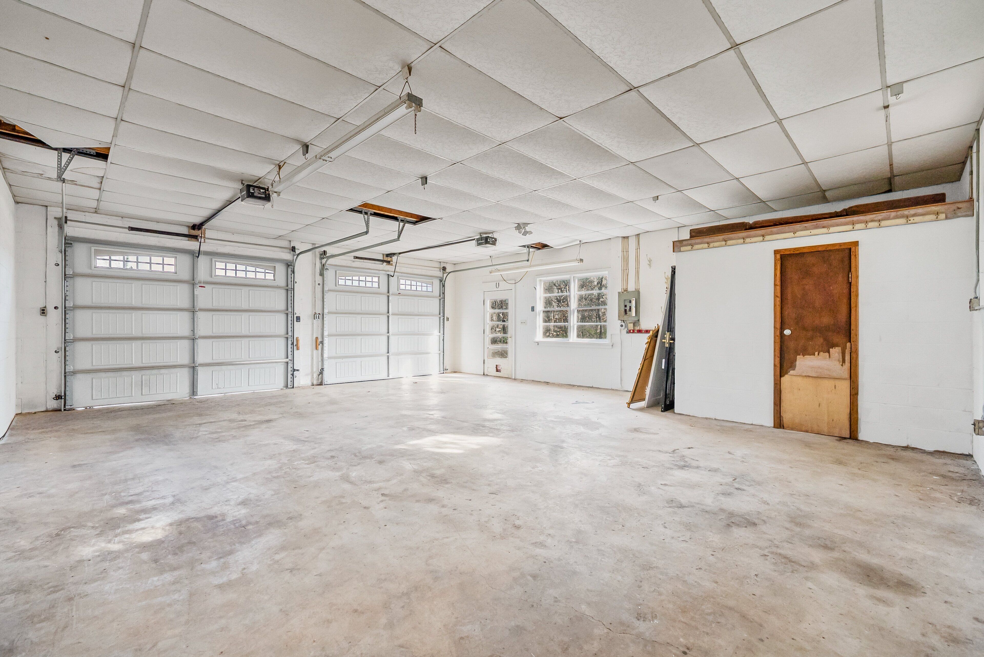A spacious garage with two large white garage doors, a concrete floor, and a wooden door on the right.